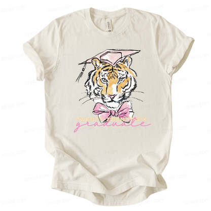 Graduation 2026 graphic t-shirt. The design features a detailed tiger illustration centered on the front, wearing a pink mortarboard cap with tassel and a large pink bow at the neck. Below the illustration, Twenty-Twenty-Six is printed in light lettering with a pink script graduate accent