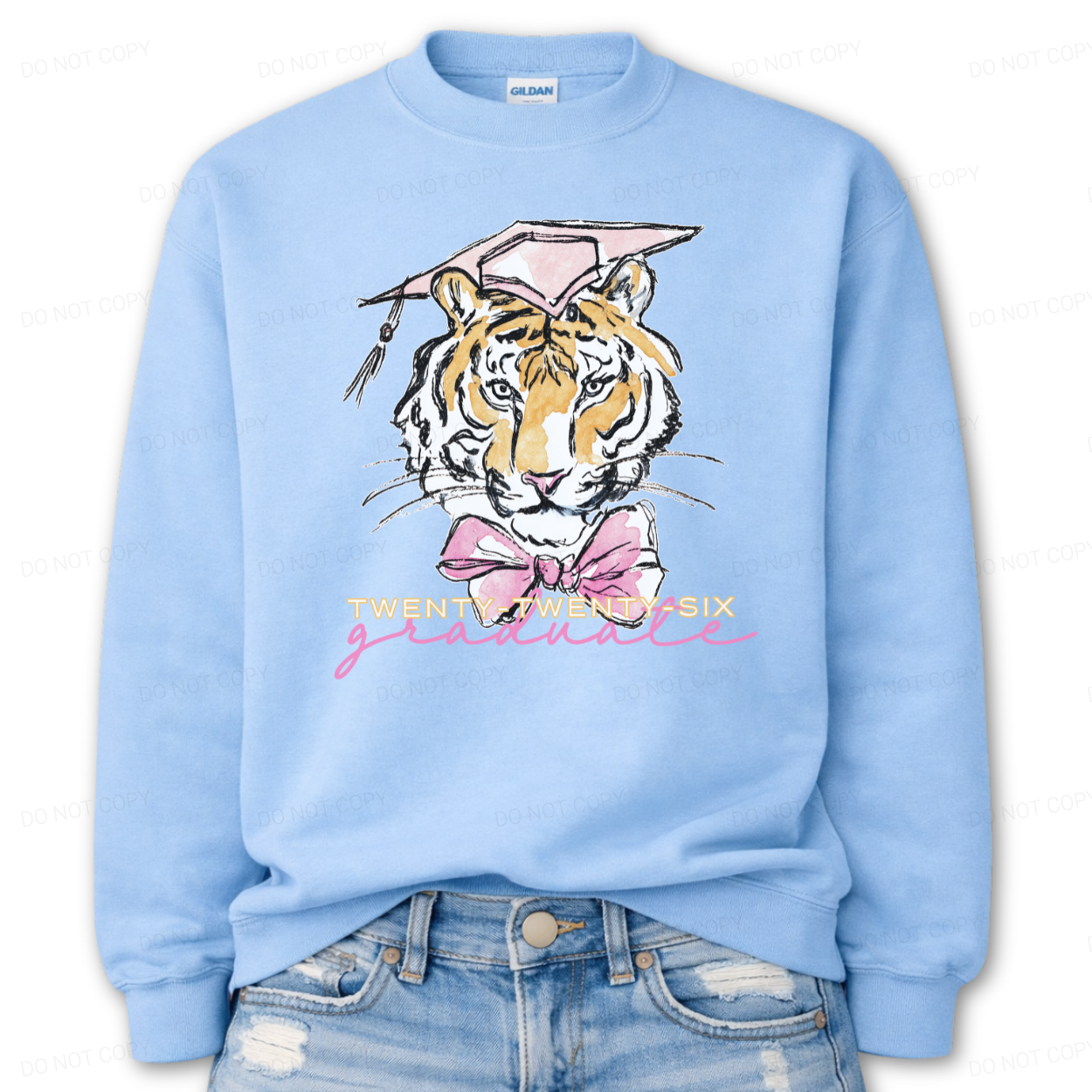 Graduation 2026 tiger graphic blue crewneck sweatshirt. The design features a detailed tiger face illustration wearing a pink graduation cap, finished with a soft pink bow at the neckline. Twenty Twenty Six graduate lettering sits below the artwork, creating a clear Class of 2026 message with a school spirit mascot look that works for both high school and college graduation customers.