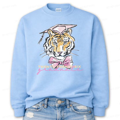 Graduation 2026 tiger graphic blue crewneck sweatshirt. The design features a detailed tiger face illustration wearing a pink graduation cap, finished with a soft pink bow at the neckline. Twenty Twenty Six graduate lettering sits below the artwork, creating a clear Class of 2026 message with a school spirit mascot look that works for both high school and college graduation customers.