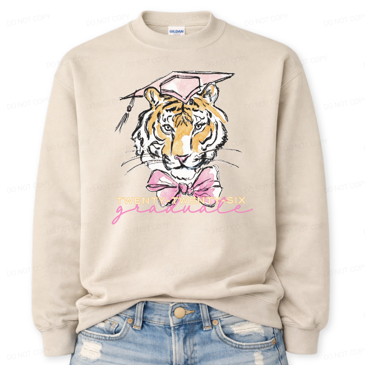 Graduation 2026 tiger graphic sand crewneck sweatshirt. The design features a detailed tiger face illustration wearing a pink graduation cap, finished with a soft pink bow at the neckline. Twenty Twenty Six graduate lettering sits below the artwork, creating a clear Class of 2026 message with a school spirit mascot look that works for both high school and college graduation customers.