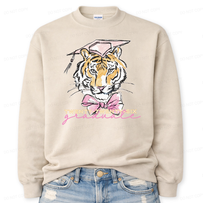 Graduation 2026 tiger graphic sand crewneck sweatshirt. The design features a detailed tiger face illustration wearing a pink graduation cap, finished with a soft pink bow at the neckline. Twenty Twenty Six graduate lettering sits below the artwork, creating a clear Class of 2026 message with a school spirit mascot look that works for both high school and college graduation customers.
