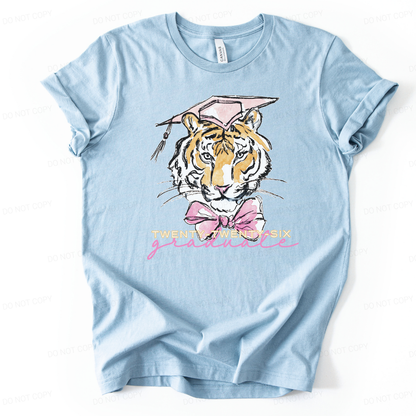 Graduation 2026 graphic t-shirt. The design features a detailed tiger illustration centered on the front, wearing a pink mortarboard cap with tassel and a large pink bow at the neck. Below the illustration, Twenty-Twenty-Six is printed in light lettering with a pink script graduate accent