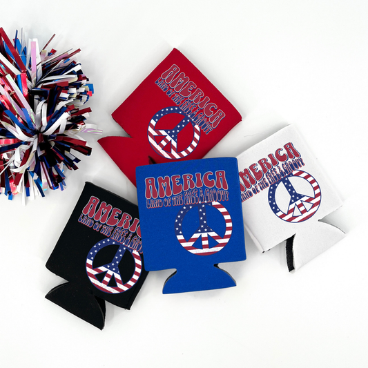 Red, Blue, Black and White collapsible can cooler displayed on a white background with a red, white, and blue metallic pom pom in the foreground. The graphic features retro lettering that reads “America Land of the Free & Groovy” above a peace sign filled with an American flag pattern. Patriotic red, white, and blue graphic printed on the front of the drink sleeve style can cooler.
