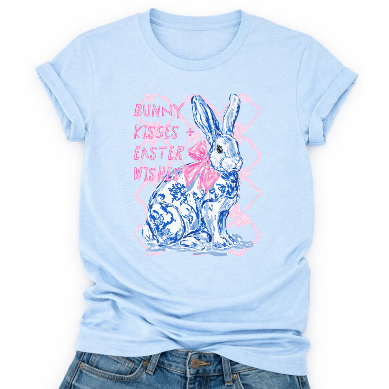 Easter spring graphic on a Bella Canvas t-shirt. The design features a hand-illustrated bunny in blue toile-style floral detailing, accented with a soft pink bow and layered over a subtle bow-pattern background. The phrase “Bunny Kisses + Easter Wishes” appears in playful pastel lettering, blending classic spring charm with boutique-ready styling.