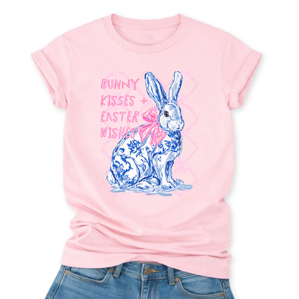 Easter spring graphic on a Bella Canvas t-shirt. The design features a hand-illustrated bunny in blue toile-style floral detailing, accented with a soft pink bow and layered over a subtle bow-pattern background. The phrase “Bunny Kisses + Easter Wishes” appears in playful pastel lettering, blending classic spring charm with boutique-ready styling.