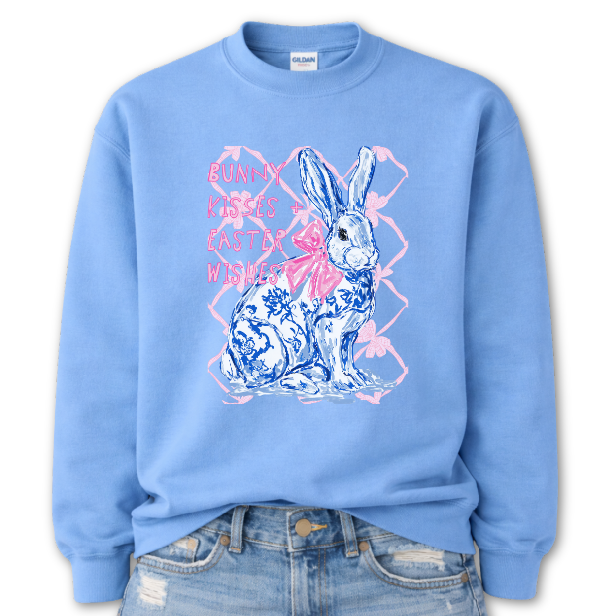 Carolina blue Bold Easter graphic on a Gildan crewneck sweatshirt. The design features a blue and white toile-style bunny with a large pink bow, layered over a soft pink lattice background and paired with the phrase “Bunny Kisses + Easter Wishes” in bright pink lettering. 