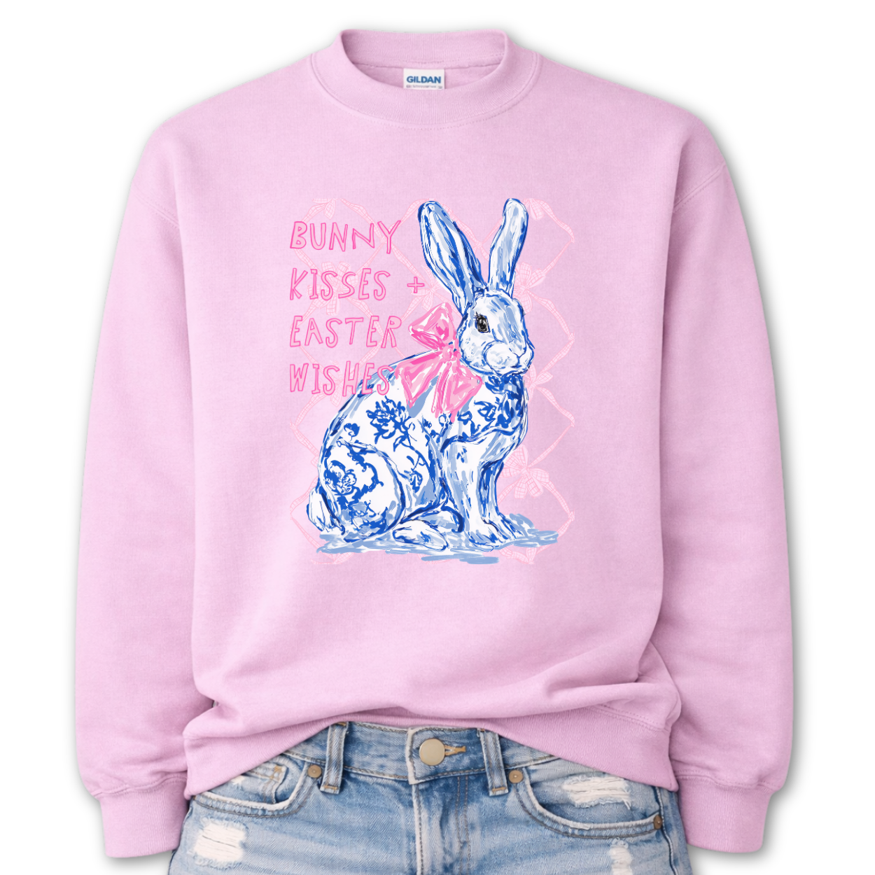 pink Bold Easter graphic on a Gildan crewneck sweatshirt. The design features a blue and white toile-style bunny with a large pink bow, layered over a soft pink lattice background and paired with the phrase “Bunny Kisses + Easter Wishes” in bright pink lettering. 