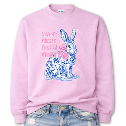 pink Bold Easter graphic on a Gildan crewneck sweatshirt. The design features a blue and white toile-style bunny with a large pink bow, layered over a soft pink lattice background and paired with the phrase “Bunny Kisses + Easter Wishes” in bright pink lettering. 