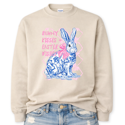 beige Bold Easter graphic on a Gildan crewneck sweatshirt. The design features a blue and white toile-style bunny with a large pink bow, layered over a soft pink lattice background and paired with the phrase “Bunny Kisses + Easter Wishes” in bright pink lettering. 