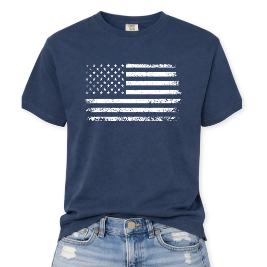 Comfort Colors Midnight t-shirt mockup with a distressed white American flag graphic, flat-lay front view with a slight front tuck into distressed denim jeans.