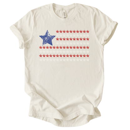 Patriotic Americana flag graphic on a cream t-shirt. The design features a distressed blue star positioned at the top left, paired with horizontal rows of red stars forming a flag-inspired layout. Soft weathered textures give the graphic a vintage, worn-in look while keeping the overall style clean and modern. The simple star-based flag design makes this an easy, everyday patriotic piece for Fourth of July, summer holidays, and Americana-themed collections.
