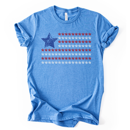Patriotic Americana flag graphic on a blue t-shirt. The design features a distressed blue star positioned at the top left, paired with horizontal rows of red stars forming a flag-inspired layout. Soft weathered textures give the graphic a vintage, worn-in look while keeping the overall style clean and modern. The simple star-based flag design makes this an easy, everyday patriotic piece for Fourth of July, summer holidays, and Americana-themed collections.