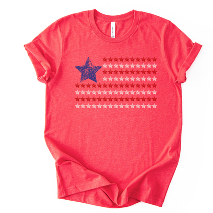 Patriotic Americana flag graphic on a red t-shirt. The design features a distressed blue star positioned at the top left, paired with horizontal rows of red stars forming a flag-inspired layout. Soft weathered textures give the graphic a vintage, worn-in look while keeping the overall style clean and modern. The simple star-based flag design makes this an easy, everyday patriotic piece for Fourth of July, summer holidays, and Americana-themed collections.