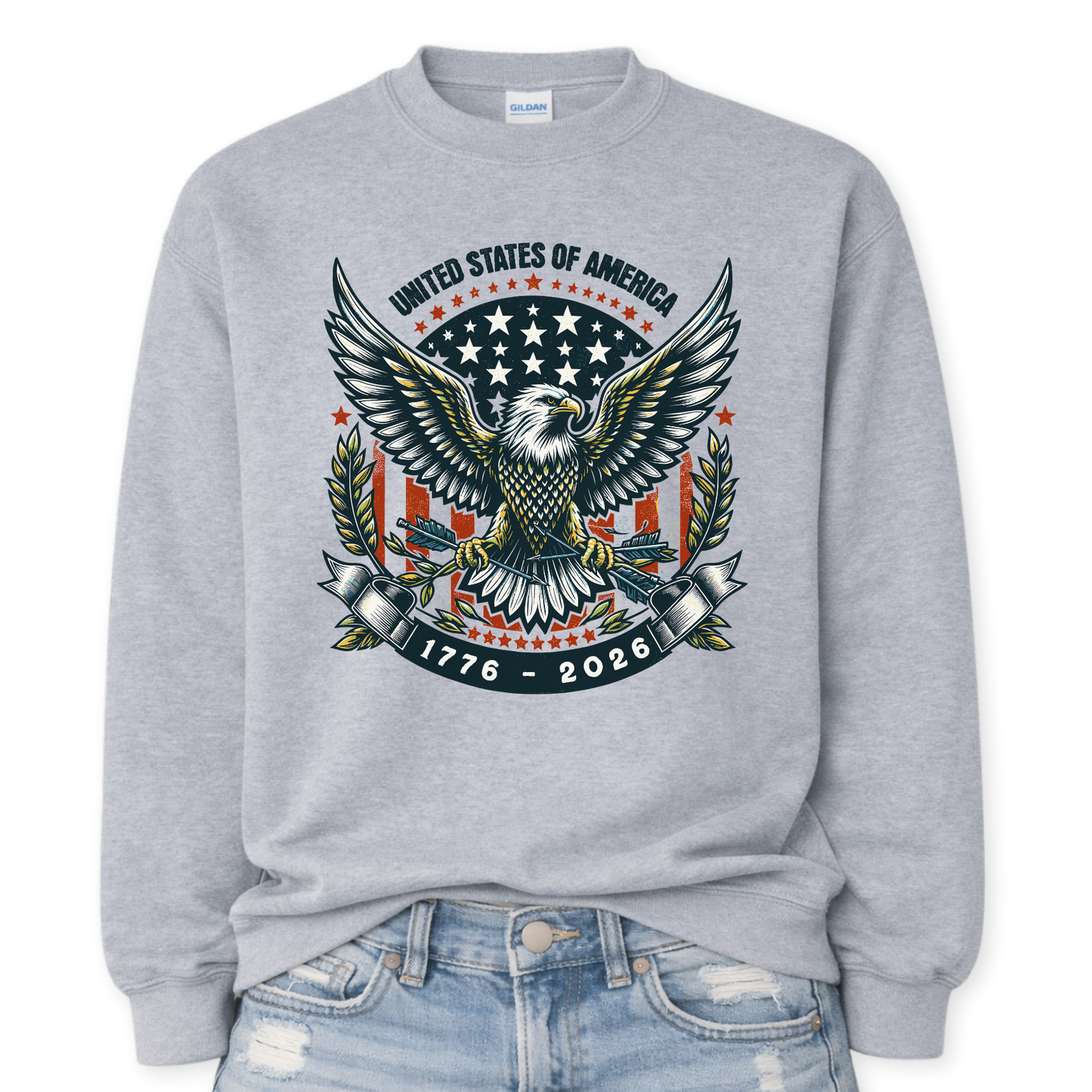 Gildan Sport Grey crewneck sweatshirt mockup with a front tuck into distressed denim, featuring a large patriotic bald eagle with wings spread, star badge backdrop, laurel branches, “United States of America,” and “1776–2026” banner graphic.