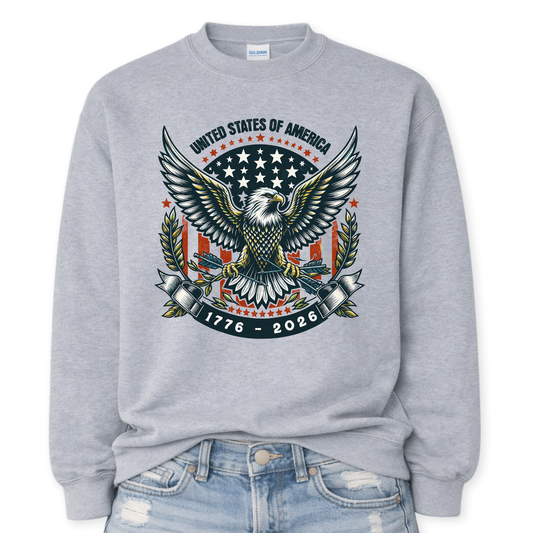 Gildan Sport Grey crewneck sweatshirt mockup with a front tuck into distressed denim, featuring a large patriotic bald eagle with wings spread, star badge backdrop, laurel branches, “United States of America,” and “1776–2026” banner graphic.