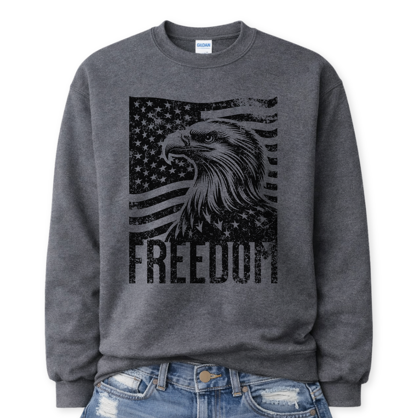 Gildan Dark Heather Grey crewneck sweatshirt mockup with a front tuck into distressed denim, featuring a black distressed American flag with a large bald eagle head overlay and “FREEDOM” block text across the bottom.