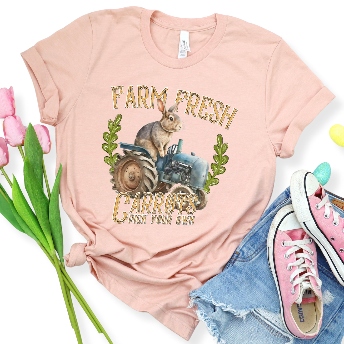 Flat lay of a Bella Canvas unisex graphic t-shirt featuring a vintage-style bunny sitting on a tractor with “Farm Fresh Carrots Pick Your Own” text, styled with jeans, sneakers, and spring decor for a playful farmhouse-inspired look.