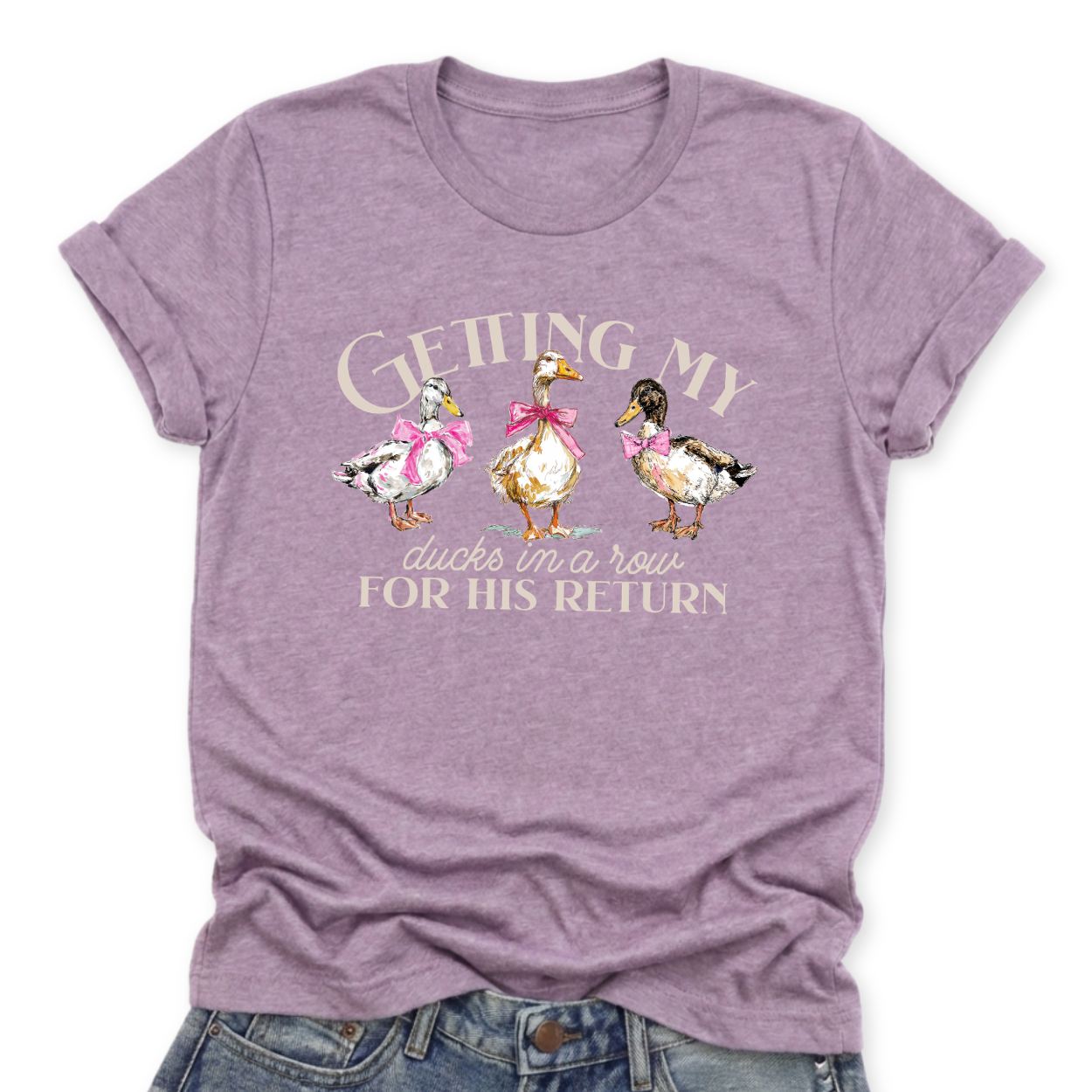 Christian Easter faith graphic on a Bella Canvas t-shirt. The design features three illustrated ducks standing side by side, each accented with soft pink bows, paired with the phrase “Getting My Ducks in a Row for His Return” in a mix of script and serif lettering. The artwork blends playful spring farmhouse style with a subtle faith-forward message