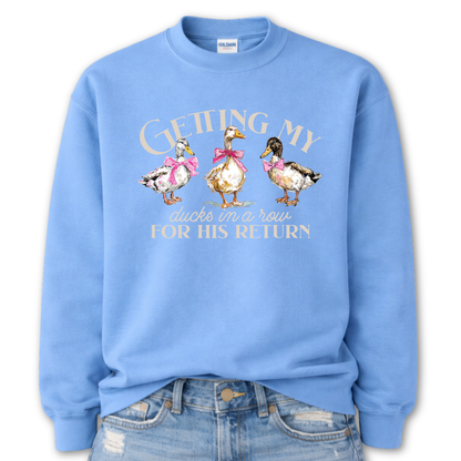 Columbia Blue Whimsical faith-based Easter graphic on a Gildan crewneck sweatshirt. The design features three hand-drawn ducks wearing pink bows, paired with the phrase “Getting My Ducks In A Row For His Return” in a soft, layered layout. The illustration style feels watercolor/sketch-inspired with gentle spring color tones.