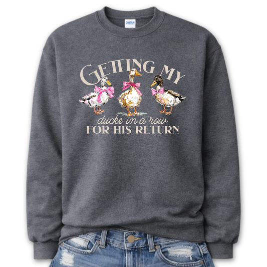 Dark Heather Gray Whimsical faith-based Easter graphic on a Gildan crewneck sweatshirt. The design features three hand-drawn ducks wearing pink bows, paired with the phrase “Getting My Ducks In A Row For His Return” in a soft, layered layout. The illustration style feels watercolor/sketch-inspired with gentle spring color tones.