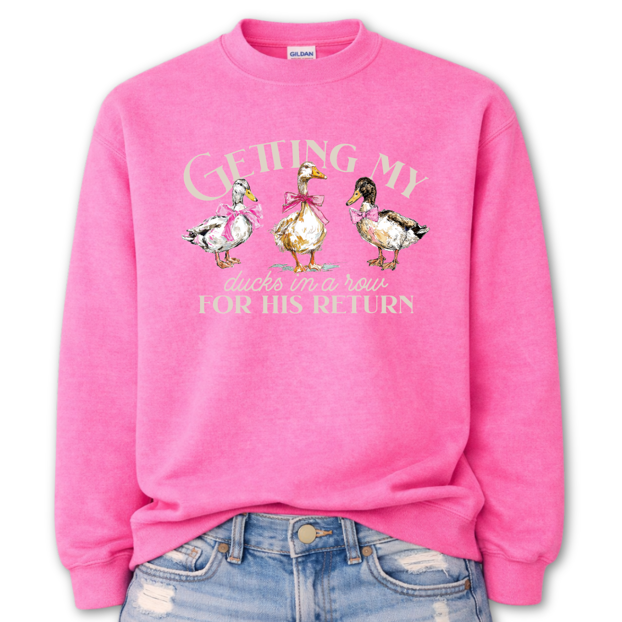 Safety Pink Whimsical faith-based Easter graphic on a Gildan crewneck sweatshirt. The design features three hand-drawn ducks wearing pink bows, paired with the phrase “Getting My Ducks In A Row For His Return” in a soft, layered layout. The illustration style feels watercolor/sketch-inspired with gentle spring color tones.