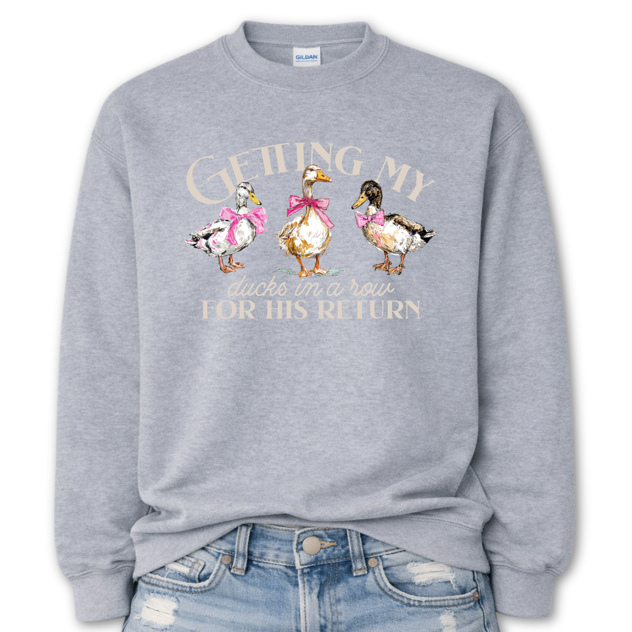 Sport Gray Whimsical faith-based Easter graphic on a Gildan crewneck sweatshirt. The design features three hand-drawn ducks wearing pink bows, paired with the phrase “Getting My Ducks In A Row For His Return” in a soft, layered layout. The illustration style feels watercolor/sketch-inspired with gentle spring color tones.