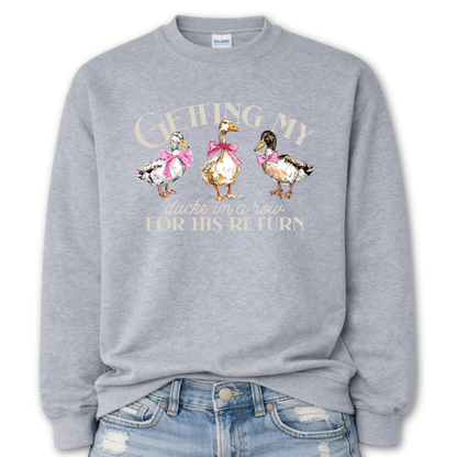 Sport Gray Whimsical faith-based Easter graphic on a Gildan crewneck sweatshirt. The design features three hand-drawn ducks wearing pink bows, paired with the phrase “Getting My Ducks In A Row For His Return” in a soft, layered layout. The illustration style feels watercolor/sketch-inspired with gentle spring color tones.
