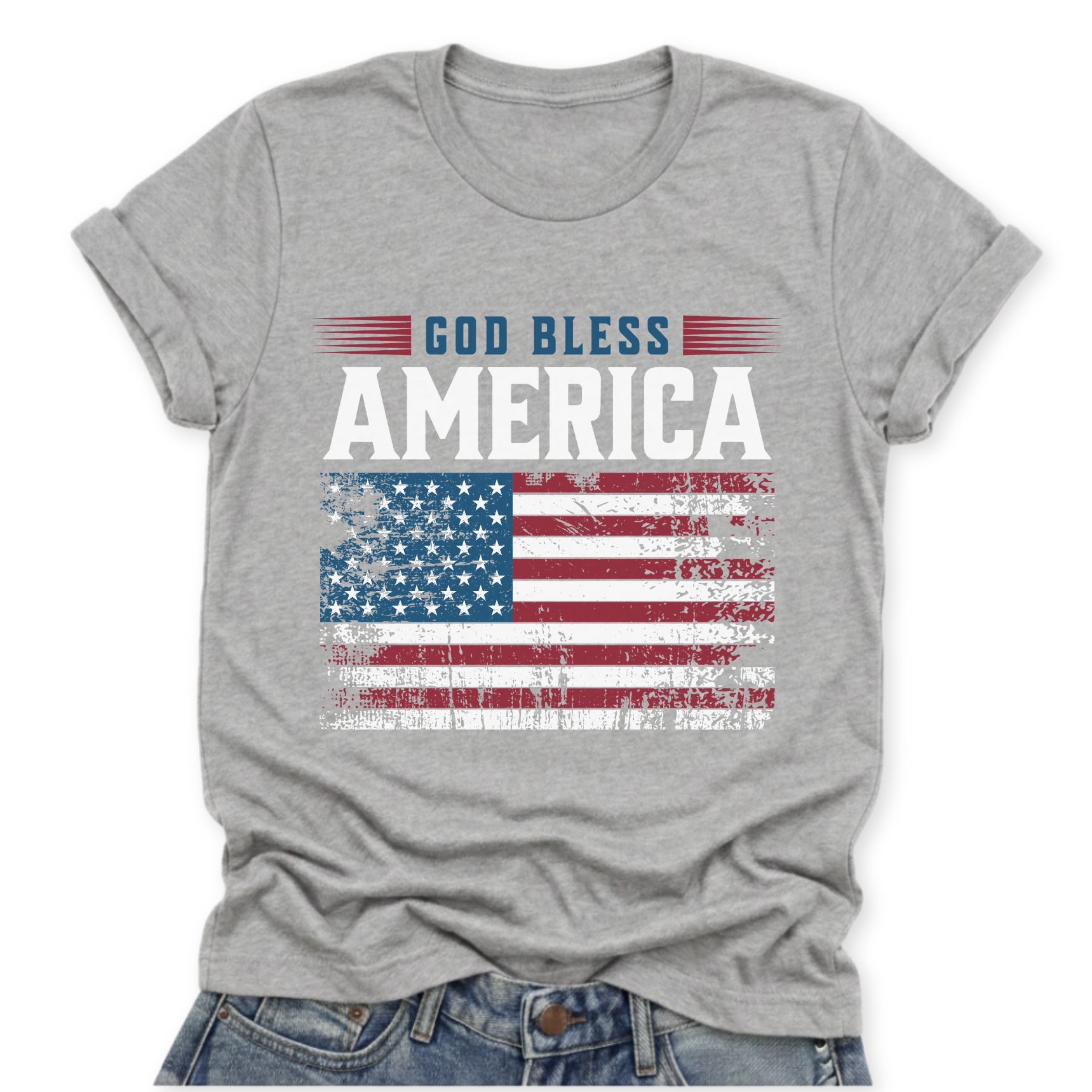 Bella Canvas athletic heather gray t-shirt with “God Bless America” text above a distressed red white and blue American flag graphic, styled in a clean flat lay with a slight front tuck into denim.