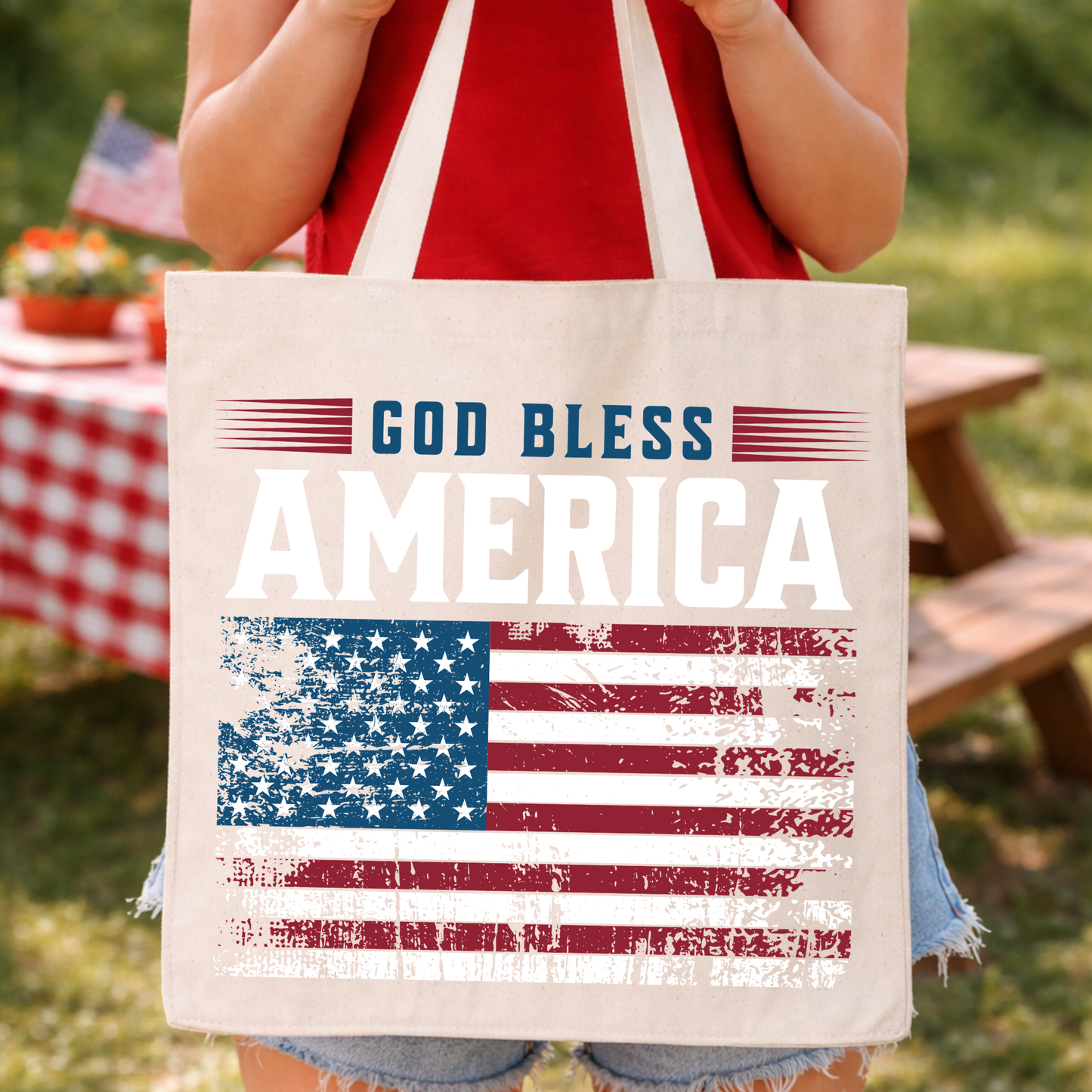 Patriotic Fourth of July faith-based graphic on a 100% cotton canvas tote bag. The design features bold “God Bless America” typography paired with a distressed American flag graphic in red, white, and blue, creating a classic Americana look.&nbsp;