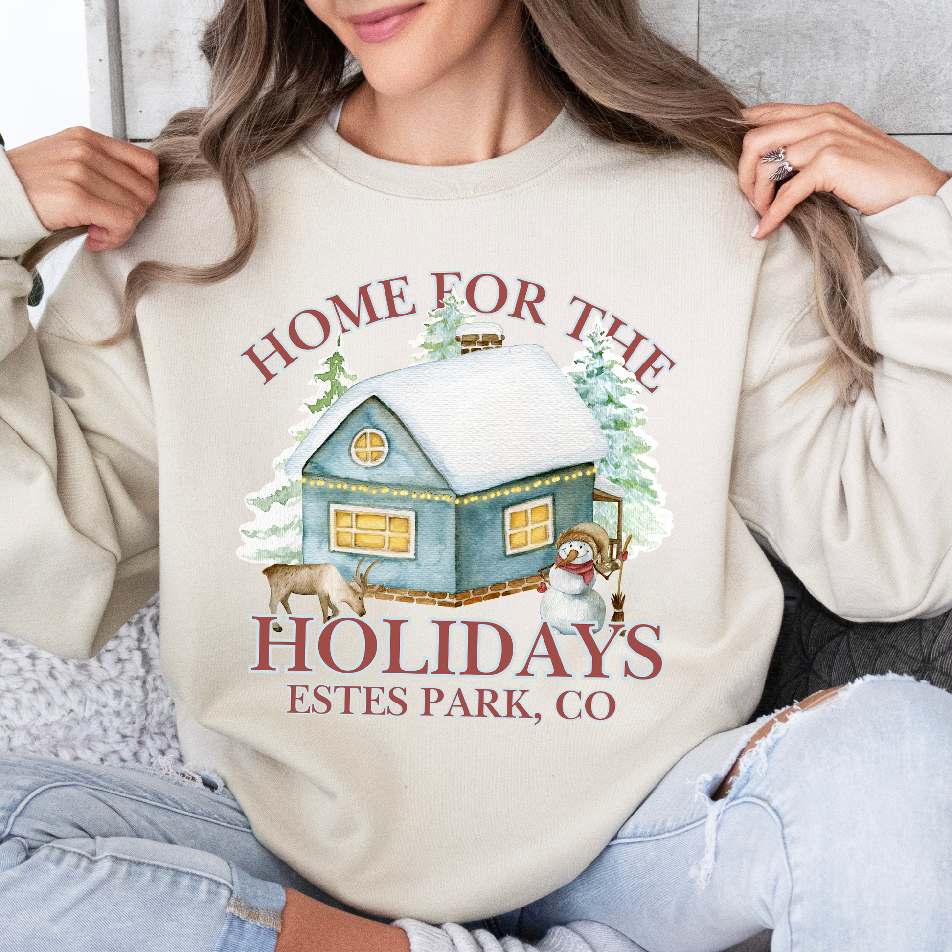 Home for the holidays beige sweatshirt with graphics of a home in the snow, trees, snowman and a deer.  customize with business name, city and state or both