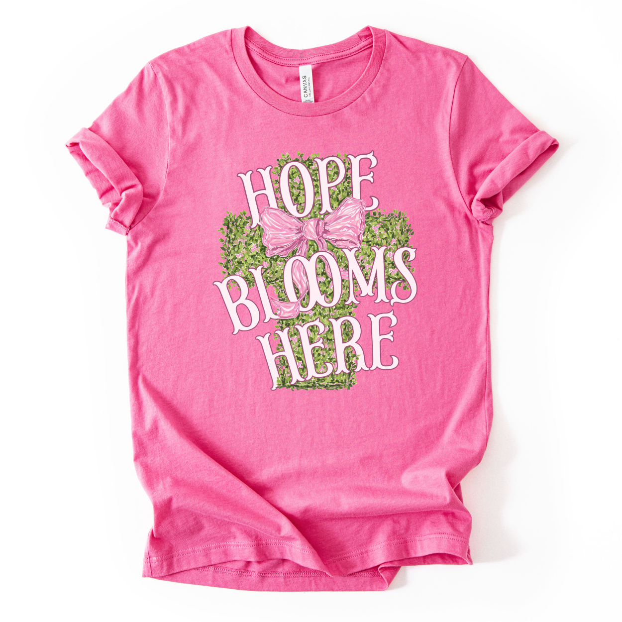 Christian spring cross graphic on a Bella Canvas t-shirt. The design features a greenery-filled cross adorned with a large pink bow, layered with the phrase “Hope Blooms Here” in bold, vintage-inspired lettering. The artwork blends Easter symbolism with soft feminine styling, creating a faith-forward seasonal piece.&nbsp;