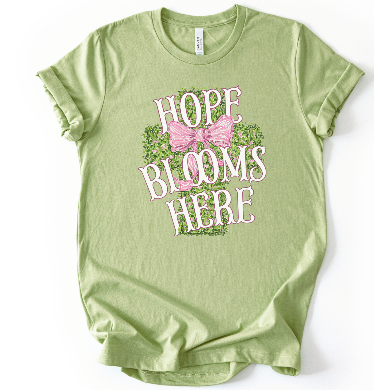 Christian spring cross graphic on a Bella Canvas t-shirt. The design features a greenery-filled cross adorned with a large pink bow, layered with the phrase “Hope Blooms Here” in bold, vintage-inspired lettering. The artwork blends Easter symbolism with soft feminine styling, creating a faith-forward seasonal piece.&nbsp;