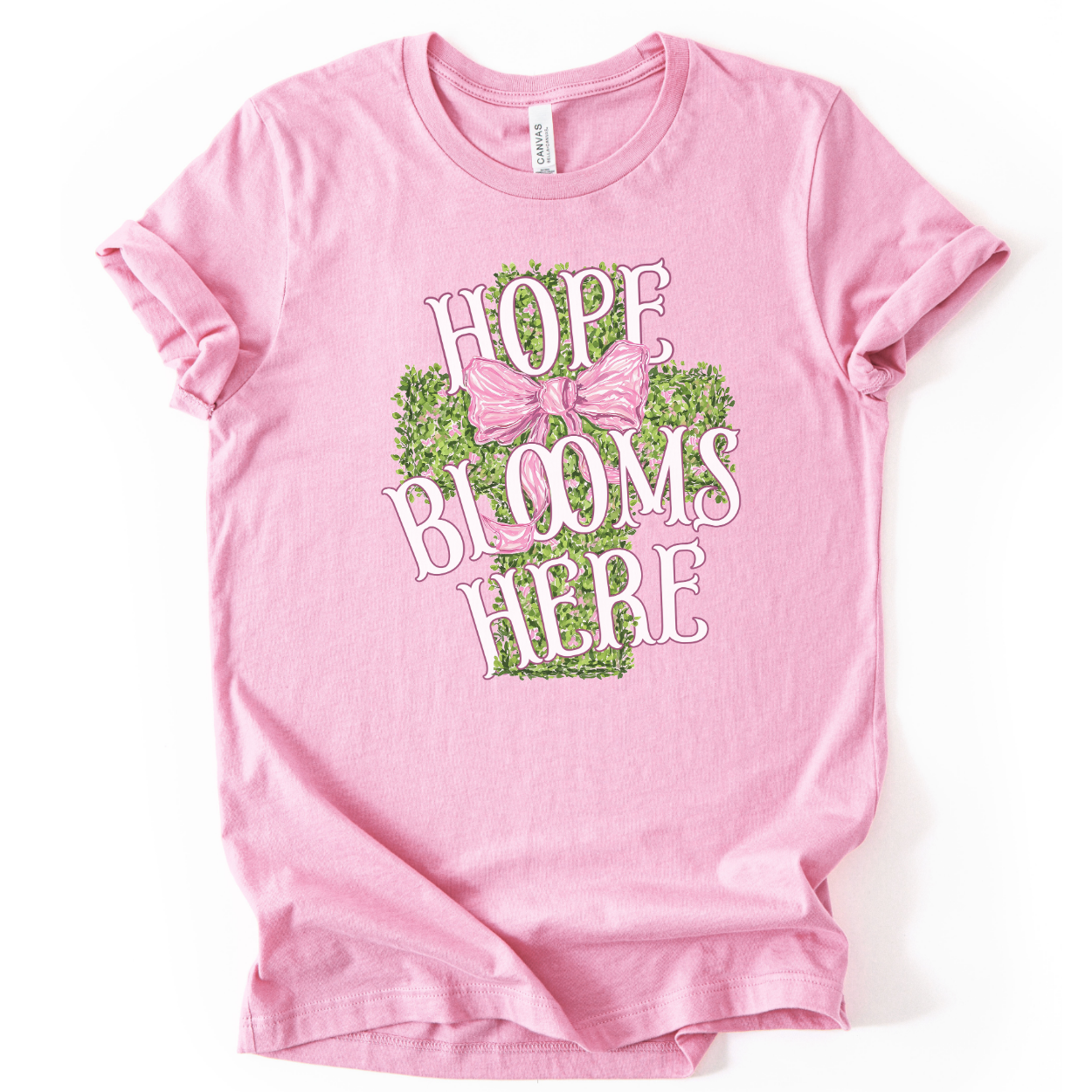 Christian spring cross graphic on a Bella Canvas t-shirt. The design features a greenery-filled cross adorned with a large pink bow, layered with the phrase “Hope Blooms Here” in bold, vintage-inspired lettering. The artwork blends Easter symbolism with soft feminine styling, creating a faith-forward seasonal piece.&nbsp;