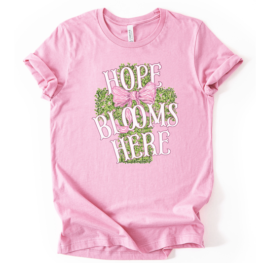 Christian spring cross graphic on a Bella Canvas t-shirt. The design features a greenery-filled cross adorned with a large pink bow, layered with the phrase “Hope Blooms Here” in bold, vintage-inspired lettering. The artwork blends Easter symbolism with soft feminine styling, creating a faith-forward seasonal piece.&nbsp;
