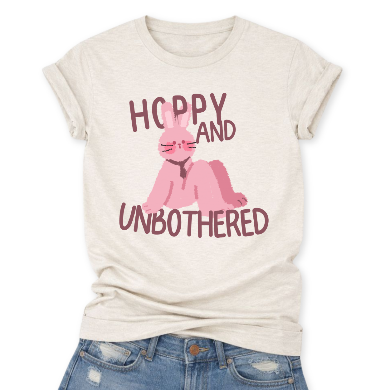 Heather Oatmeal Playful Easter graphic on a Bella Canvas t-shirt. The design features a relaxed pink bunny illustration paired with the bold phrase “Hoppy and Unbothered” in modern, oversized lettering. The artwork blends trendy humor with soft seasonal styling, making it a fun spring statement piece.
