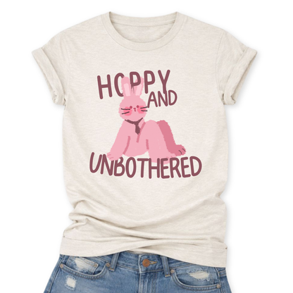 Heather Oatmeal Playful Easter graphic on a Bella Canvas t-shirt. The design features a relaxed pink bunny illustration paired with the bold phrase “Hoppy and Unbothered” in modern, oversized lettering. The artwork blends trendy humor with soft seasonal styling, making it a fun spring statement piece.