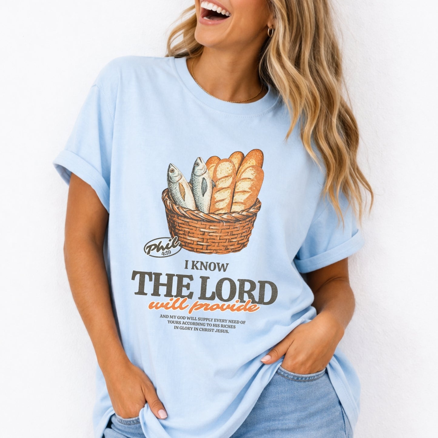 Person wearing a light blue t-shirt with a graphic design and text.  I know the Lord will provide featuring a basket of bread and fish referencing bible verse Philipians 4:19