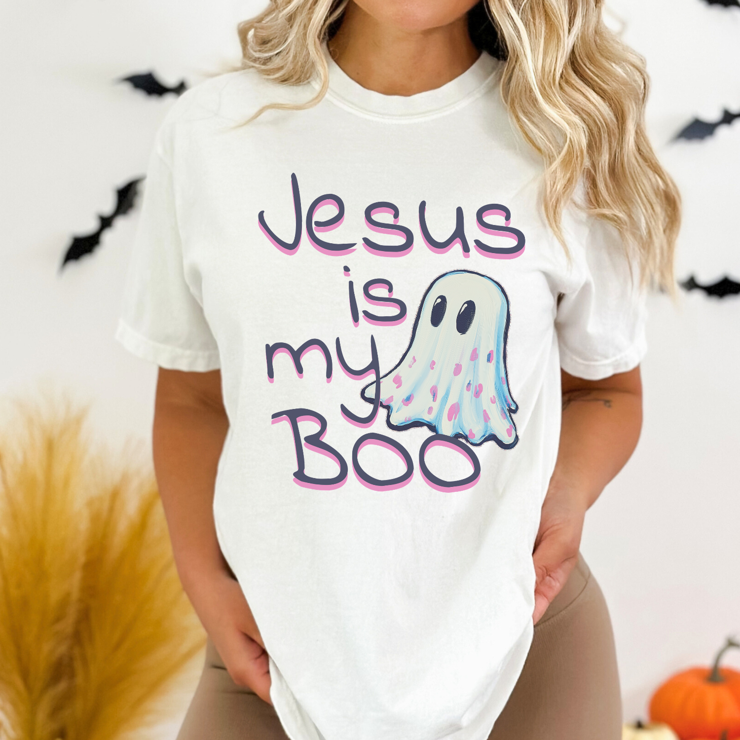 A woman wearing a white graphic t-shirt with the text 'Jesus is my Boo' in purple and pink bubble letters next to a cute blue and pink ghost illustration. The background is a white wall decorated with black bat silhouettes and pampas grass.