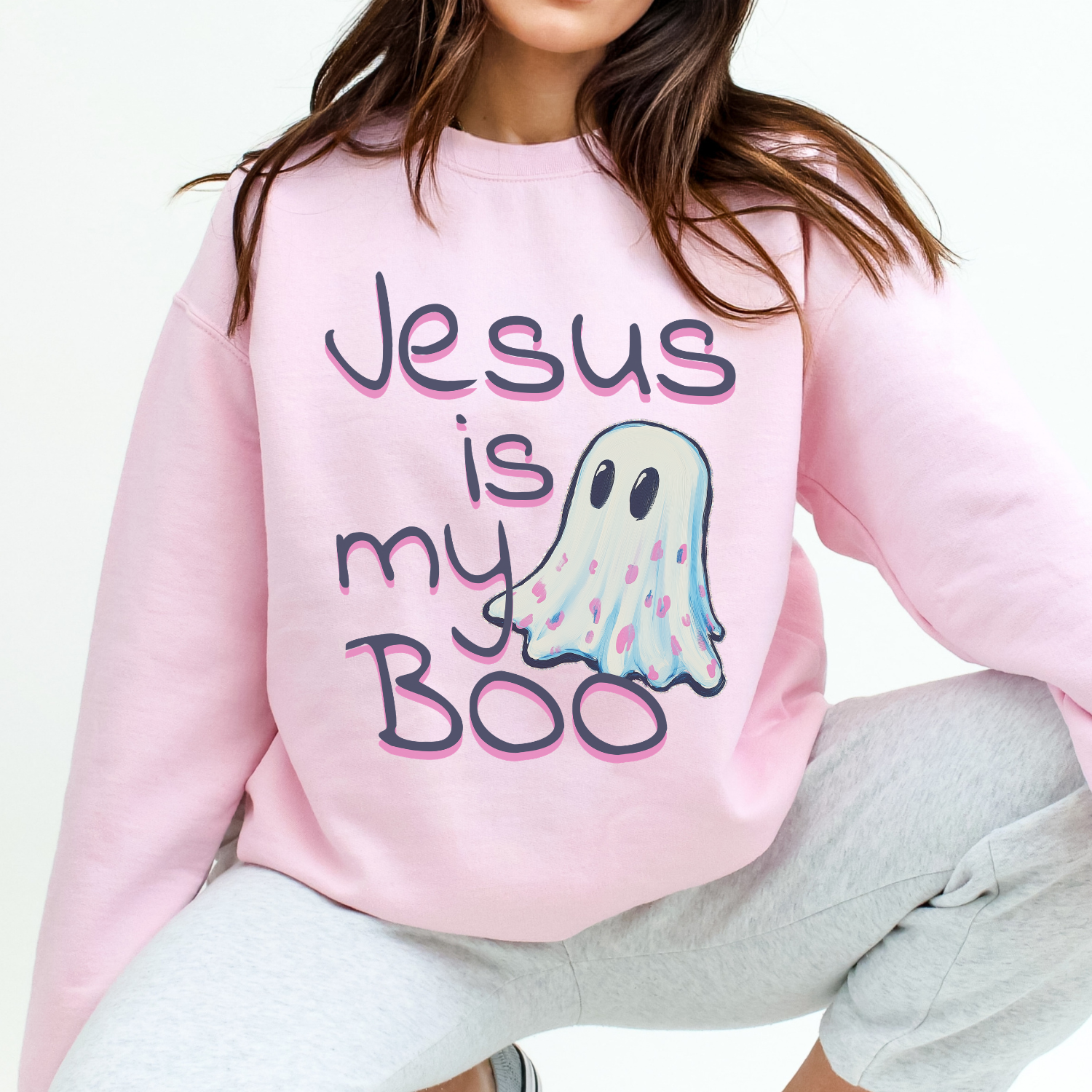 A person wearing a light pink crewneck sweatshirt with a graphic that reads 'Jesus is my Boo' in dark purple lettering with a pink outline, next to a cute pastel ghost illustration. The person is also wearing light gray sweatpants against a clean white background.
