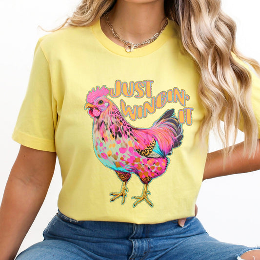 Person wearing a yellow t-shirt with a colorful rooster graphic and 'Just Wingin' It' text.