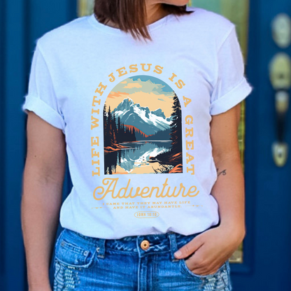 A person wearing a white graphic t-shirt with text and a mountainous landscape design. Scripture text Life with Jesus is a Great Adventure John 10:10