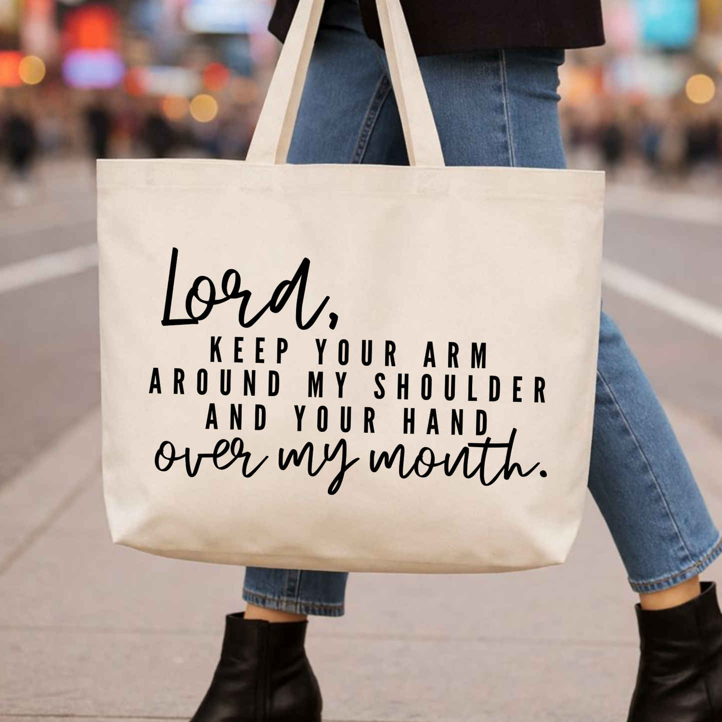 Natural cotton canvas tote bag with black mixed-font lettering reading “Lord, keep your arm around my shoulder and your hand over my mouth,” carried by a woman wearing jeans on a blurred city street background; large reusable canvas tote with a faith-based, humorous message.