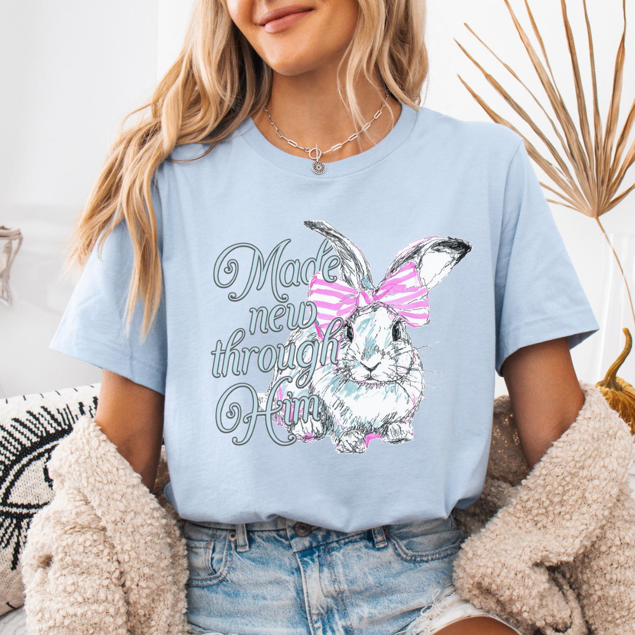 Christian Easter resurrection graphic on a Bella Canvas t-shirt. The design features a hand-sketched bunny accented with a pink bow and soft pastel shading, paired with the phrase “Made New Through Him” in flowing, feminine lettering. The artwork highlights the renewal message of Christ and the hope of Easter, blending soft spring styling with a strong faith-forward statement.