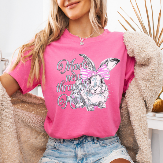 Christian Easter resurrection graphic on a Bella Canvas t-shirt. The design features a hand-sketched bunny accented with a pink bow and soft pastel shading, paired with the phrase “Made New Through Him” in flowing, feminine lettering. The artwork highlights the renewal message of Christ and the hope of Easter, blending soft spring styling with a strong faith-forward statement.