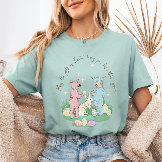 Easter seasonal graphic on a Bella Canvas t-shirt. The design features a vintage-inspired illustration of two children dressed in bunny outfits gathered around a rabbit with pastel Easter eggs and soft greenery. The phrase “May the gifts of Easter bring you hope that grows” curves above the artwork in delicate script lettering, creating a nostalgic, heartfelt spring message.