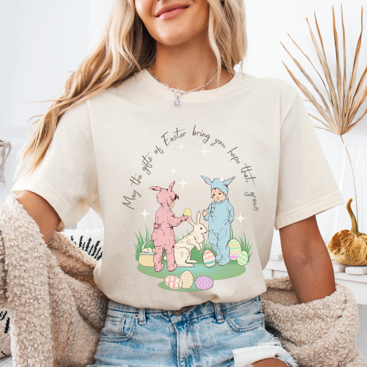Easter seasonal graphic on a Bella Canvas t-shirt. The design features a vintage-inspired illustration of two children dressed in bunny outfits gathered around a rabbit with pastel Easter eggs and soft greenery. The phrase “May the gifts of Easter bring you hope that grows” curves above the artwork in delicate script lettering, creating a nostalgic, heartfelt spring message.