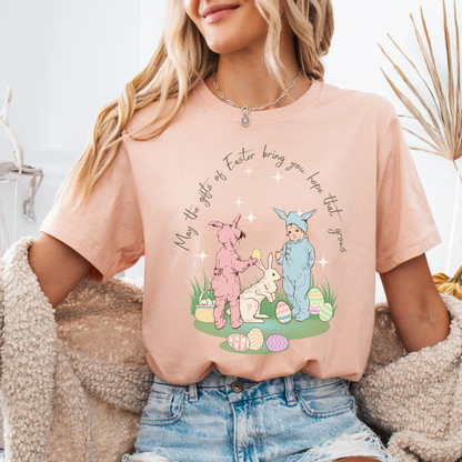 Easter seasonal graphic on a Bella Canvas t-shirt. The design features a vintage-inspired illustration of two children dressed in bunny outfits gathered around a rabbit with pastel Easter eggs and soft greenery. The phrase “May the gifts of Easter bring you hope that grows” curves above the artwork in delicate script lettering, creating a nostalgic, heartfelt spring message.