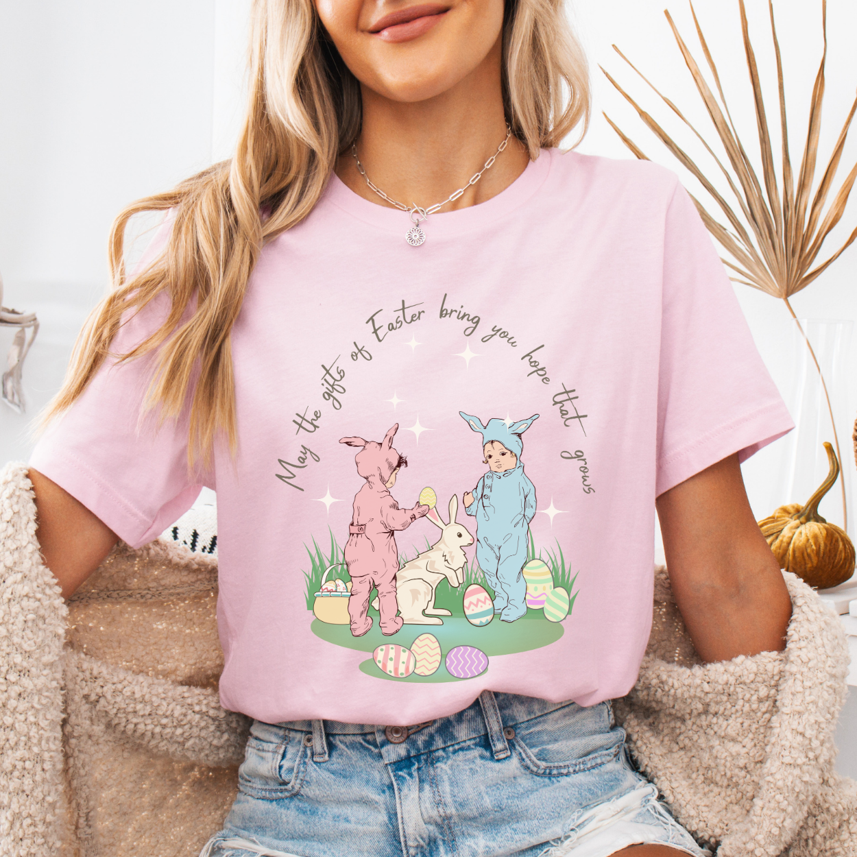 Easter seasonal graphic on a Bella Canvas t-shirt. The design features a vintage-inspired illustration of two children dressed in bunny outfits gathered around a rabbit with pastel Easter eggs and soft greenery. The phrase “May the gifts of Easter bring you hope that grows” curves above the artwork in delicate script lettering, creating a nostalgic, heartfelt spring message.