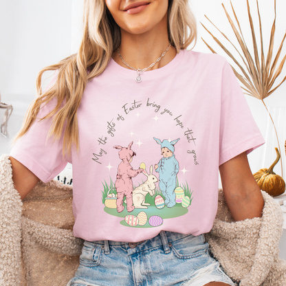 Easter seasonal graphic on a Bella Canvas t-shirt. The design features a vintage-inspired illustration of two children dressed in bunny outfits gathered around a rabbit with pastel Easter eggs and soft greenery. The phrase “May the gifts of Easter bring you hope that grows” curves above the artwork in delicate script lettering, creating a nostalgic, heartfelt spring message.