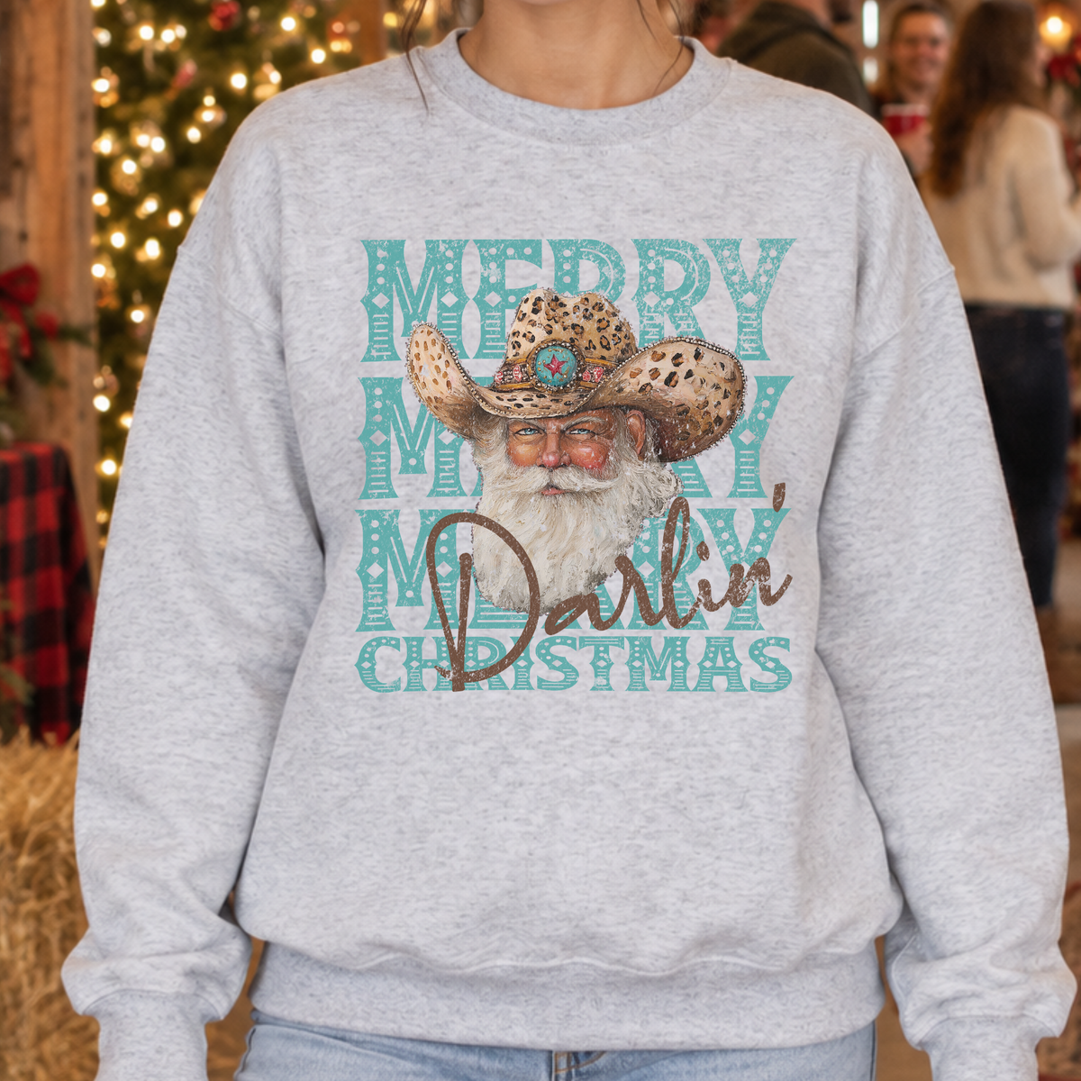 A person wearing a light heather gray sweatshirt with a festive graphic that reads 'Merry Merry Merry Christmas' in teal, featuring a cowboy hat and Santa's beard, with 'Darlin'' in brown script. The background shows a softly blurred holiday scene with warm lights and people.